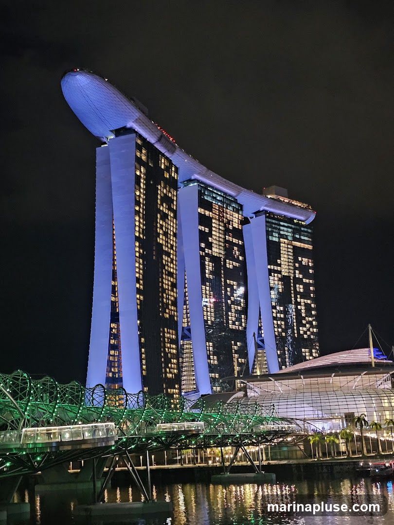A nighttime view of the Marina Bay Sands hotel in Singapore, with its iconic three towers topped by a ship-like structure. The building is illuminated with blue and yellow lights. In the foreground, the Helix Bridge, a pedestrian bridge, is lit with green lights and reflects in the water below. The complex appears to be a major tourist attraction and architectural landmark.
