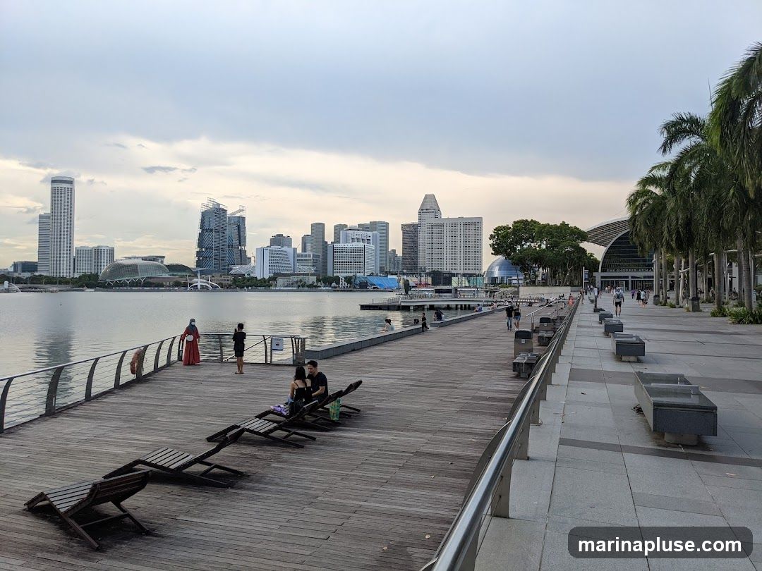 A wide wooden promenade runs along a body of water, with several people relaxing on lounge chairs and walking. In the background, a modern city skyline rises, featuring tall buildings and distinctive architectural structures. Palm trees line the right side of the promenade, leading towards a large building with a curved roof. The sky is overcast, suggesting a cloudy day.