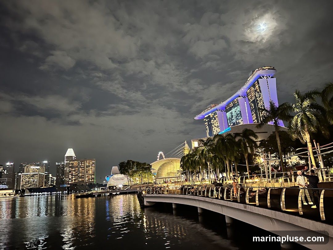 A nighttime view of a cityscape, featuring the iconic Marina Bay Sands hotel illuminated with purple lights. The hotel is surrounded by palm trees and a cloudy sky with a visible moon. In the foreground, a waterfront promenade is lined with people, and the water reflects the city lights. Other buildings and structures are visible in the background, creating a vibrant urban scene.