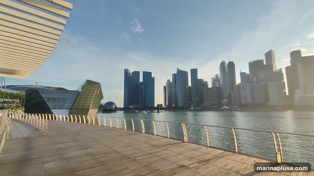 A wide-angle view of the Marina Bay area in Singapore, featuring the distinctive architecture of Marina Bay Sands on the left, with its geometric glass structure and a domed building behind it. The iconic Singapore skyline with numerous skyscrapers forms the backdrop across the water. A wooden boardwalk with metal railings occupies the foreground, leading the eye towards the cityscape. The sky is clear with a few wispy clouds, and the sun casts long shadows, suggesting it is either early morning or late afternoon.