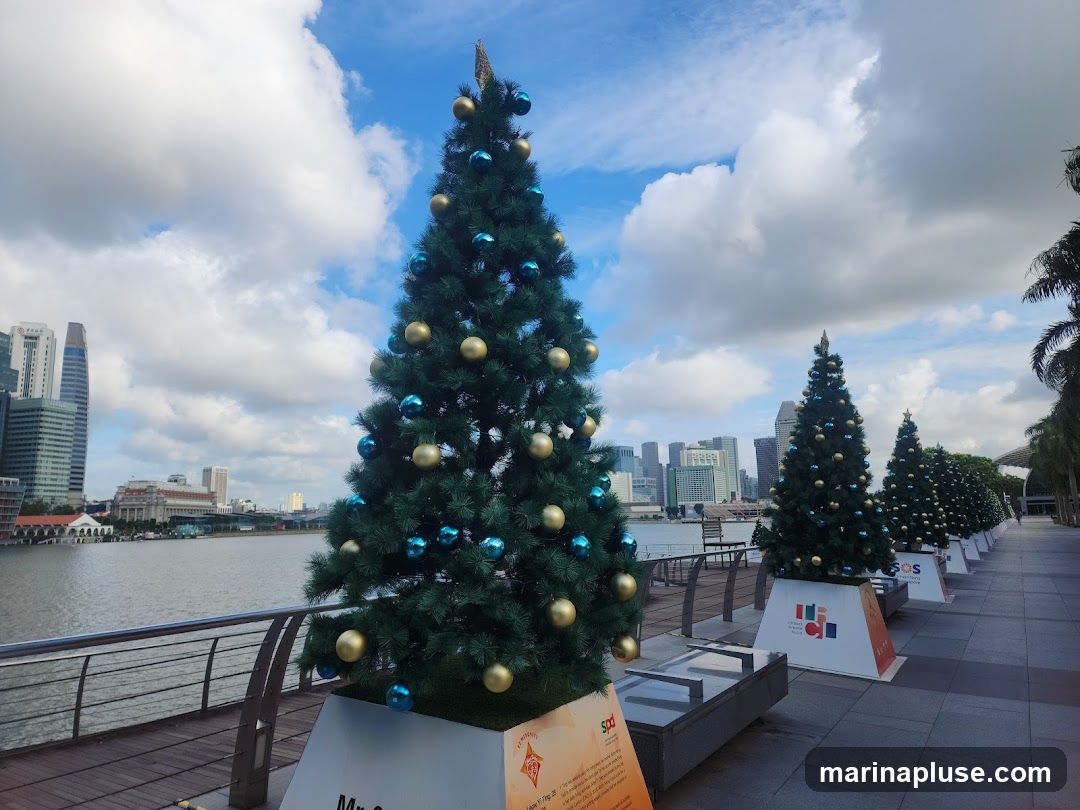 A row of decorated Christmas trees stands along a waterfront promenade, with a city skyline in the background. The trees are adorned with blue and gold ornaments. The sky is partly cloudy.