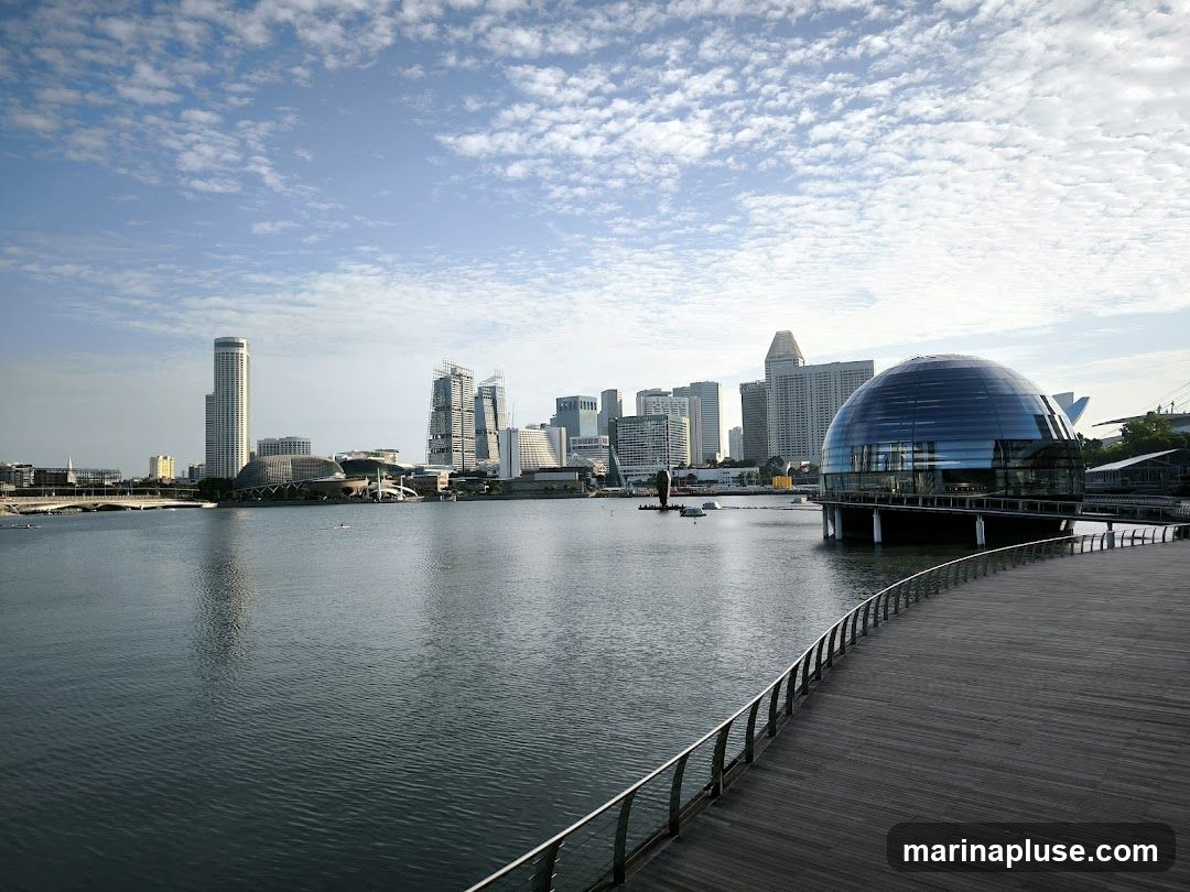 A wide wooden promenade curves into the foreground, bordered by a metal railing. Beyond the promenade, a calm body of water reflects the sky and the distant city skyline. The skyline features various modern skyscrapers, including a tall, slender building on the left and a cluster of taller buildings in the center. To the right, a distinctive spherical building with a glass facade stands on stilts over the water. The sky is partly cloudy with scattered white clouds against a light blue backdrop. A few small boats are visible on the water.