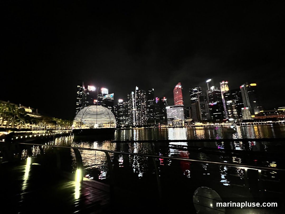 A nighttime view of a modern cityscape with a prominent spherical building illuminated from within, situated on the waterfront. The building's glass facade reflects the surrounding lights. In the background, a cluster of skyscrapers glows with various colored lights, creating a vibrant urban panorama. The water in the foreground mirrors the city lights, adding depth and atmosphere to the scene. Palm trees line the waterfront to the left of the spherical structure.