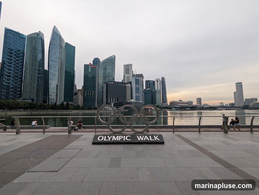 A wide shot of the Olympic rings sculpture with the words "OLYMPIC WALK" below it, set against a backdrop of a modern city skyline and a body of water. Several people are visible sitting and walking along the waterfront promenade. The sky is overcast, suggesting early morning or late afternoon light.