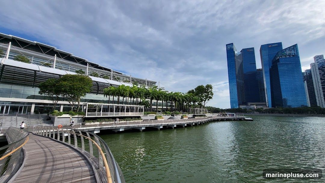 A wide shot of the Marina Bay waterfront in Singapore. On the left is a modern building with a sloped roof and glass facade, surrounded by trees and palm trees. A curved wooden walkway leads into the foreground. In the background, several tall, blue glass skyscrapers, including the DBS and HSBC buildings, rise against a cloudy sky. The water of the bay is calm and reflects the sky.