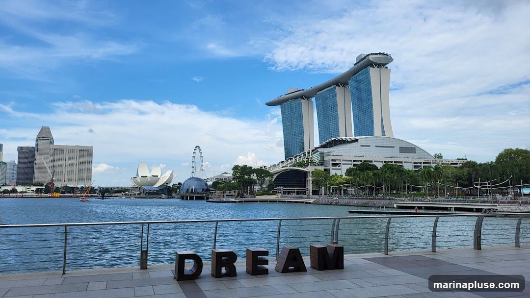 A wide shot of the Singapore skyline featuring the iconic Marina Bay Sands hotel, the ArtScience Museum, and the Singapore Flyer. The foreground shows a waterfront promenade with large wooden letters spelling out "DREAM". The water in Marina Bay is calm, reflecting the blue sky and white clouds.