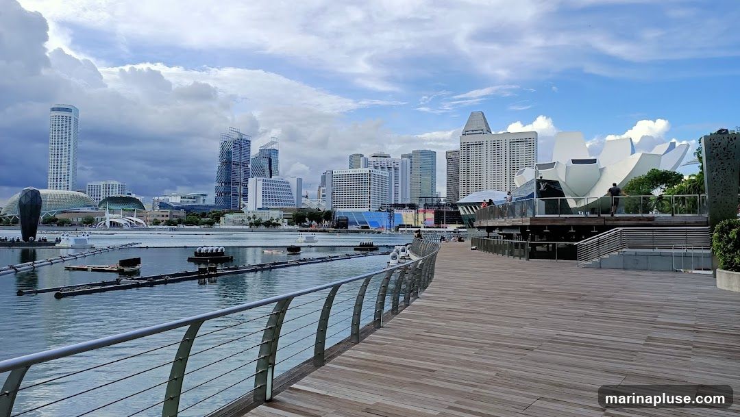 A wide wooden promenade runs along the edge of a body of water, with a railing separating it from the water. In the distance, a modern city skyline is visible, featuring numerous skyscrapers of varying heights and architectural styles. The sky is partly cloudy with dramatic cloud formations. On the right side of the promenade, a distinctive white, lotus-shaped building with a Louis Vuitton logo is partially visible. People are seen walking on the promenade and near the building.