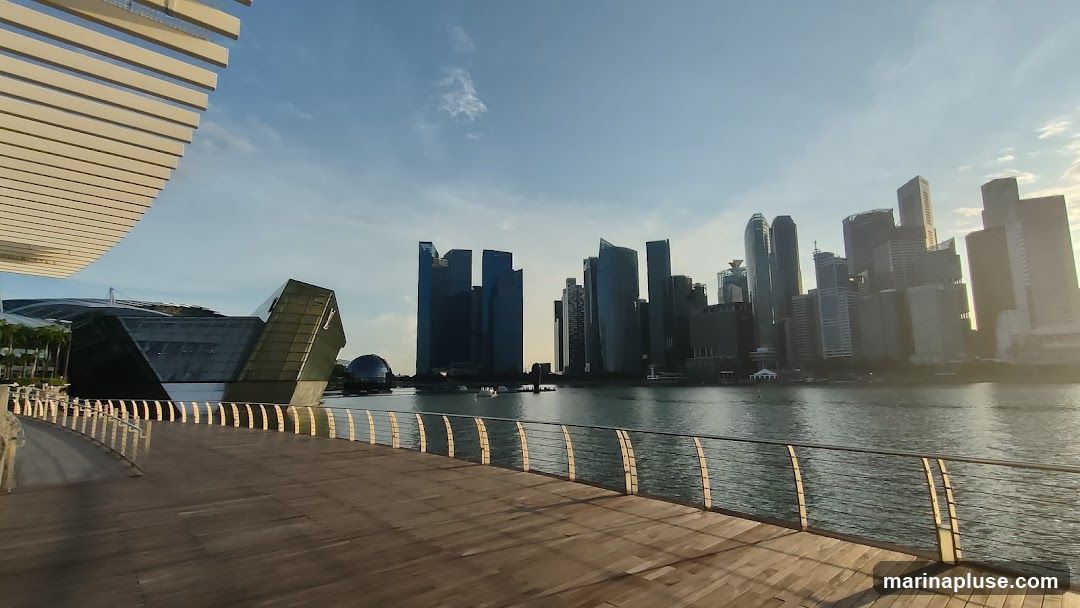 A wide wooden promenade with metal railings curves along the foreground, leading towards a body of water. Across the water, a modern city skyline rises, dominated by numerous skyscrapers of varying heights and architectural styles. On the left side of the image, a distinctive building with a geometric, glass-paneled facade is visible, with a large, curved, slatted roof structure extending over the promenade. The sky above is a clear blue with a few wispy clouds, and the sun casts long shadows across the promenade, indicating it is either early morning or late afternoon.
