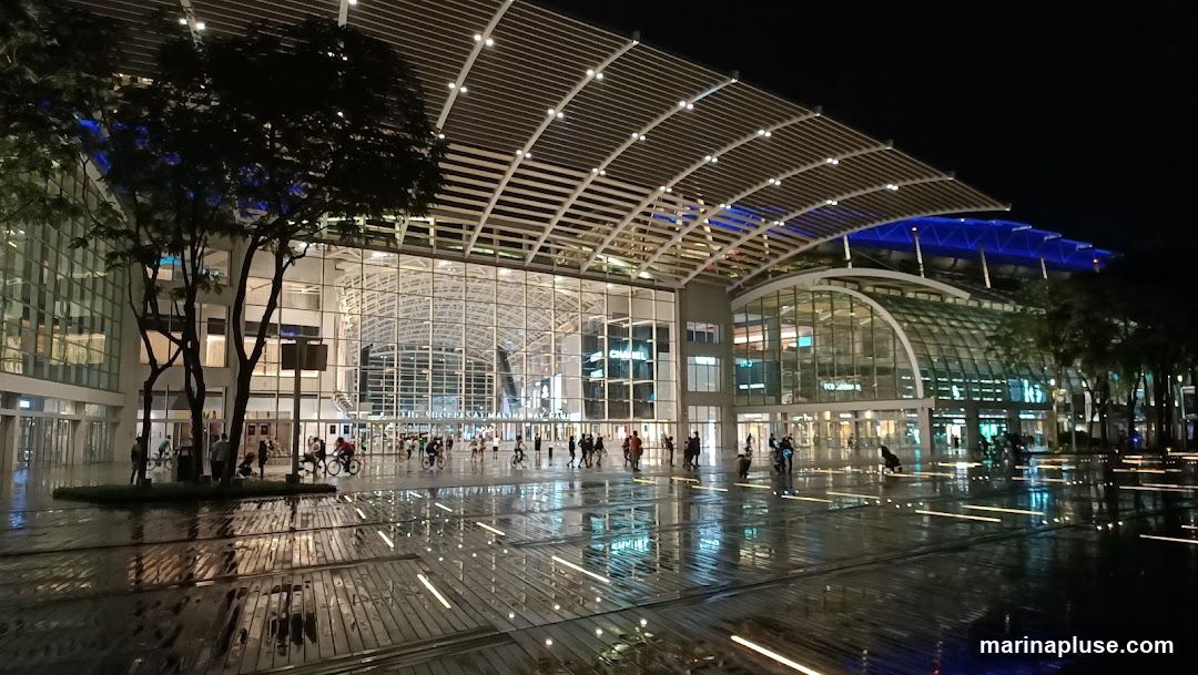 A wide shot of a modern building at night, with a large, arched, glass-covered entrance. The facade is illuminated by lights, and the wet ground reflects the lights and the building. People are visible walking and cycling in front of the building. The architecture features a distinctive, slatted roof structure.