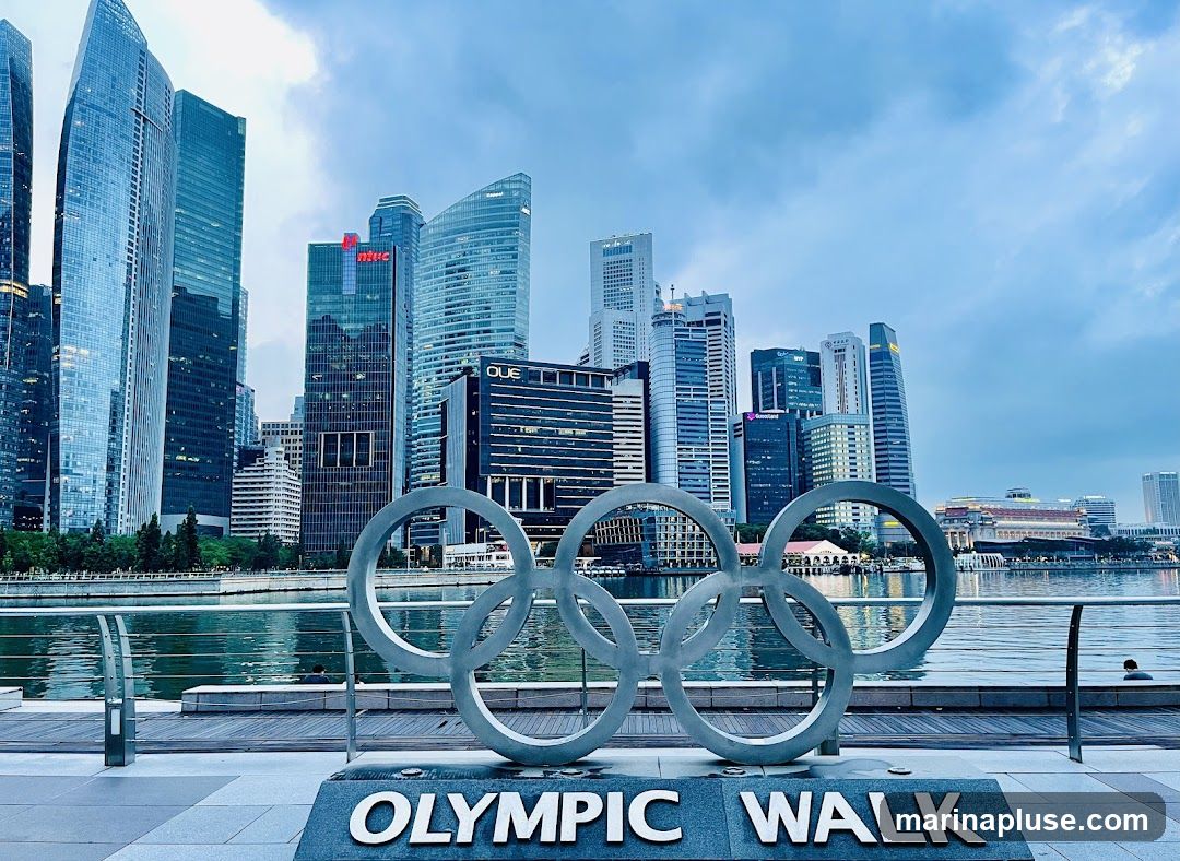 A large sculpture of the Olympic rings stands in the foreground, with the words "OLYMPIC WALK" visible on its base. Behind the sculpture, a modern city skyline with numerous skyscrapers rises against a cloudy sky. A body of water, likely a river or bay, separates the sculpture from the buildings. The architecture of the buildings is varied, with some featuring sleek, curved designs and others appearing more traditional. The overall scene suggests an urban waterfront area with a connection to the Olympic Games.