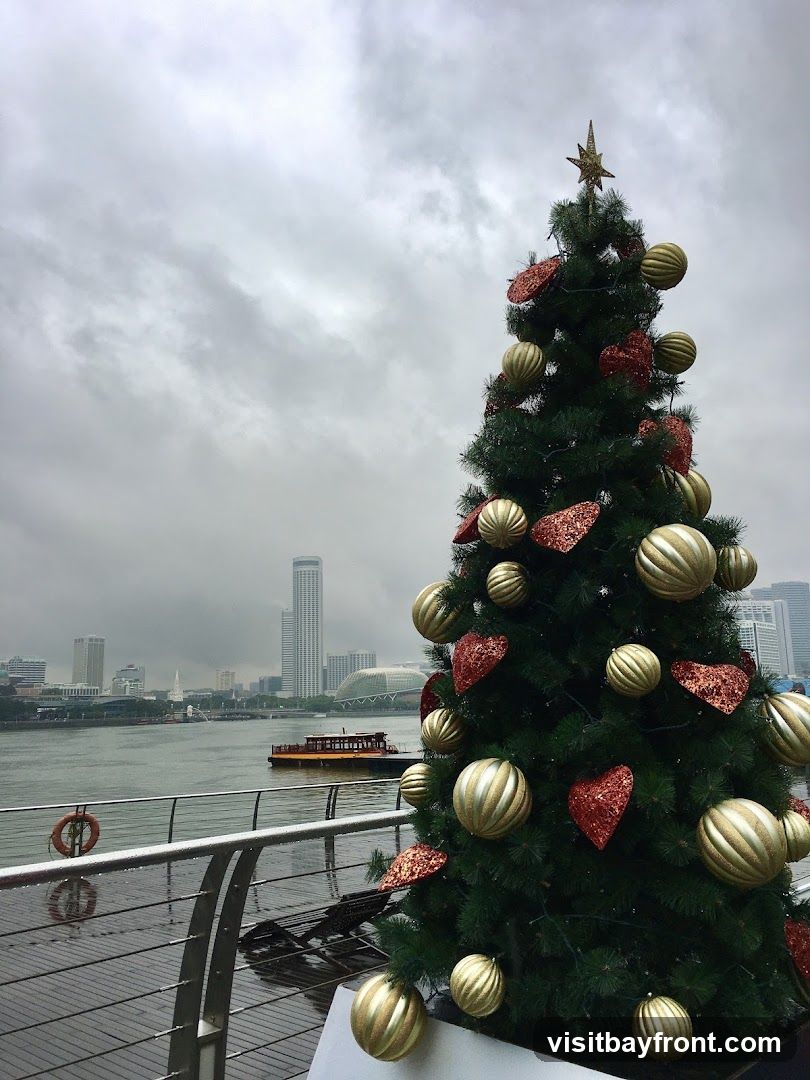 A decorated Christmas tree stands on a wooden deck next to a railing, with a body of water and a city skyline in the background. The tree is adorned with gold and red ornaments, including large spherical baubles and heart-shaped glitter decorations. A star tops the tree. The water is calm, and a boat is visible in the distance. The sky is overcast and cloudy.