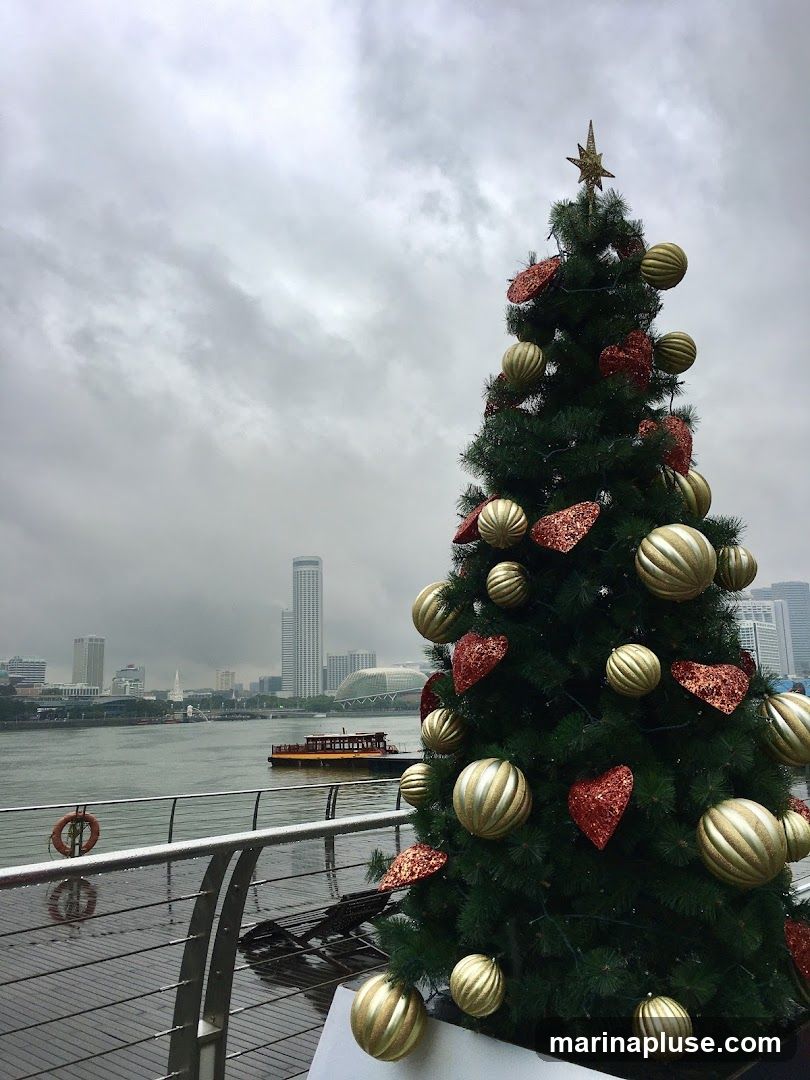 A decorated Christmas tree stands on a wooden deck next to a railing, with a body of water and a city skyline in the background. The tree is adorned with gold and red ornaments, including large spherical baubles and heart-shaped glitter decorations. A star tops the tree. The water is calm, and a boat is visible in the distance. The sky is overcast and cloudy.