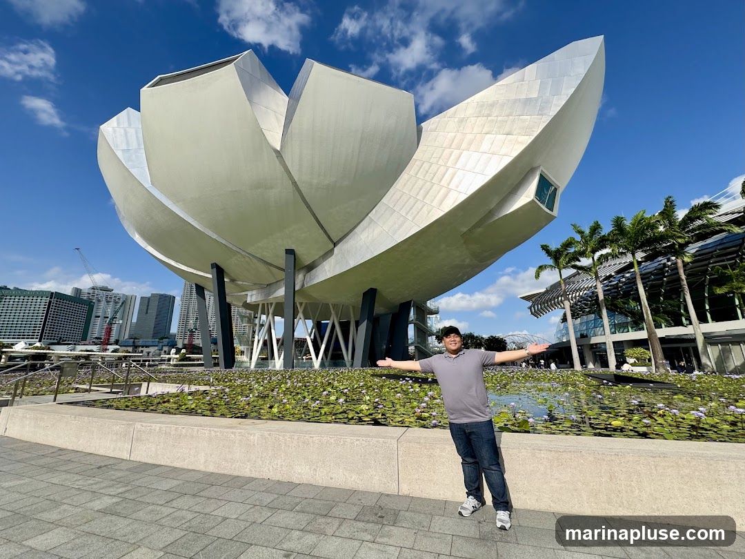 A man in a grey polo shirt and blue jeans stands with his arms outstretched in front of a large, modern, lotus-shaped building. The building has a metallic, petal-like exterior and is supported by dark pillars. In the foreground, there is a pond filled with water lilies. In the background, a cityscape with tall buildings and palm trees can be seen under a clear blue sky with scattered clouds.