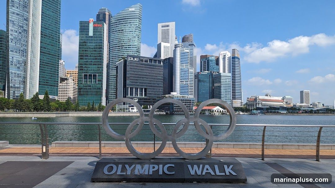 A large sculpture of the Olympic rings stands prominently in the foreground, with the words "OLYMPIC WALK" inscribed on its base. Behind the sculpture, a wide body of water separates the viewer from a dense cityscape featuring numerous modern skyscrapers of varying heights and architectural styles. The sky above is a clear blue with scattered white clouds. The scene appears to be a waterfront promenade in a bustling urban environment.