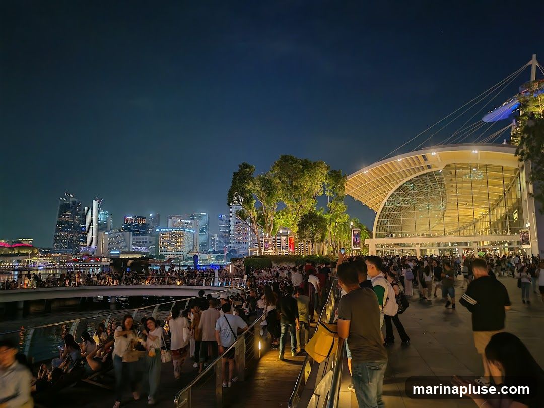 A bustling waterfront scene at night, with a large crowd of people gathered. In the background, a modern building with a distinctive curved roof is illuminated, and beyond that, a city skyline with numerous skyscrapers is visible. The area appears to be a popular public space, possibly a promenade or entertainment district.