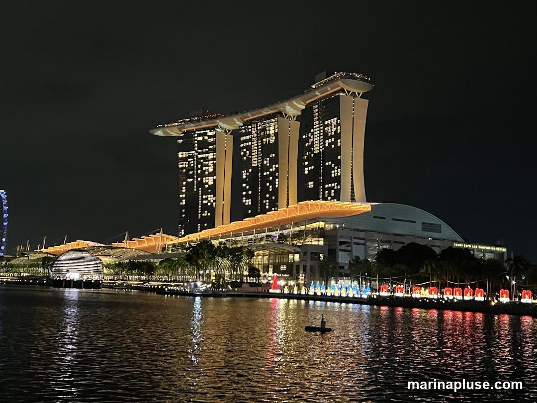 A nighttime view of the Marina Bay Sands complex in Singapore, with its iconic three towers and connecting SkyPark. The building is illuminated, and its lights are reflected in the calm water of the bay. Along the waterfront, there are decorative lights, possibly for a festive occasion, in red and blue. A small boat is visible on the water.