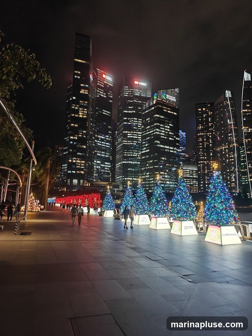 A nighttime view of a city skyline with illuminated skyscrapers. In the foreground, a paved walkway is lined with decorated Christmas trees and festive lights. People are seen strolling along the walkway. The buildings display corporate logos such as DBS, HSBC, and Standard Chartered.
