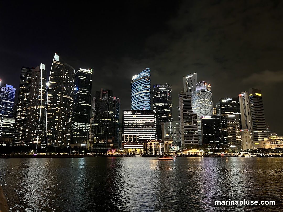 A nighttime view of a modern city skyline with numerous illuminated skyscrapers reflected in the water below. A boat is visible on the water.