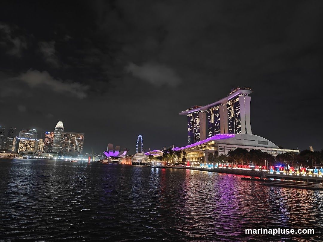 A nighttime view of the Singapore skyline, featuring the iconic Marina Bay Sands hotel illuminated with purple lights. The cityscape includes other tall buildings and a Ferris wheel, all reflected in the dark water of the bay. The waterfront is lined with trees and decorative lights.