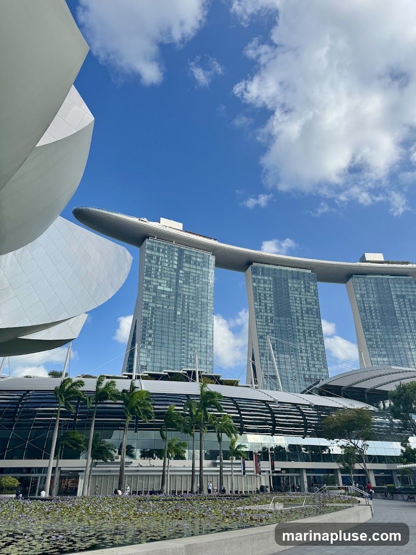 A wide shot of the Marina Bay Sands complex in Singapore on a sunny day with a blue sky and scattered clouds. The iconic three towers are visible, connected by the SkyPark. In the foreground, there is a large body of water with lily pads and a walkway. Palm trees line the front of the lower building, which has a modern, glass-fronted facade.
