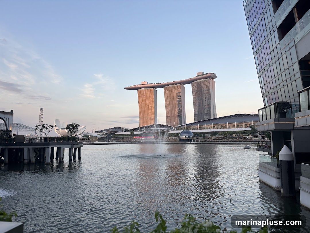 A wide shot of the Marina Bay Sands hotel and its iconic three towers, connected by a rooftop deck, stands prominently against a clear blue sky. In the foreground, a calm body of water reflects the surrounding buildings and a fountain sprays water upwards. To the right, a modern glass-fronted building juts into the frame, its facade catching the light. On the left, a Ferris wheel and other structures are visible, suggesting a vibrant urban landscape. The scene is bathed in the soft light of either dawn or dusk.