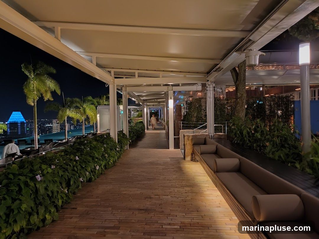 A long, covered walkway with brick paving and lush greenery lining one side. Comfortable seating areas with plush cushions are visible along the other side. In the distance, a cityscape with illuminated buildings and palm trees can be seen under a night sky.