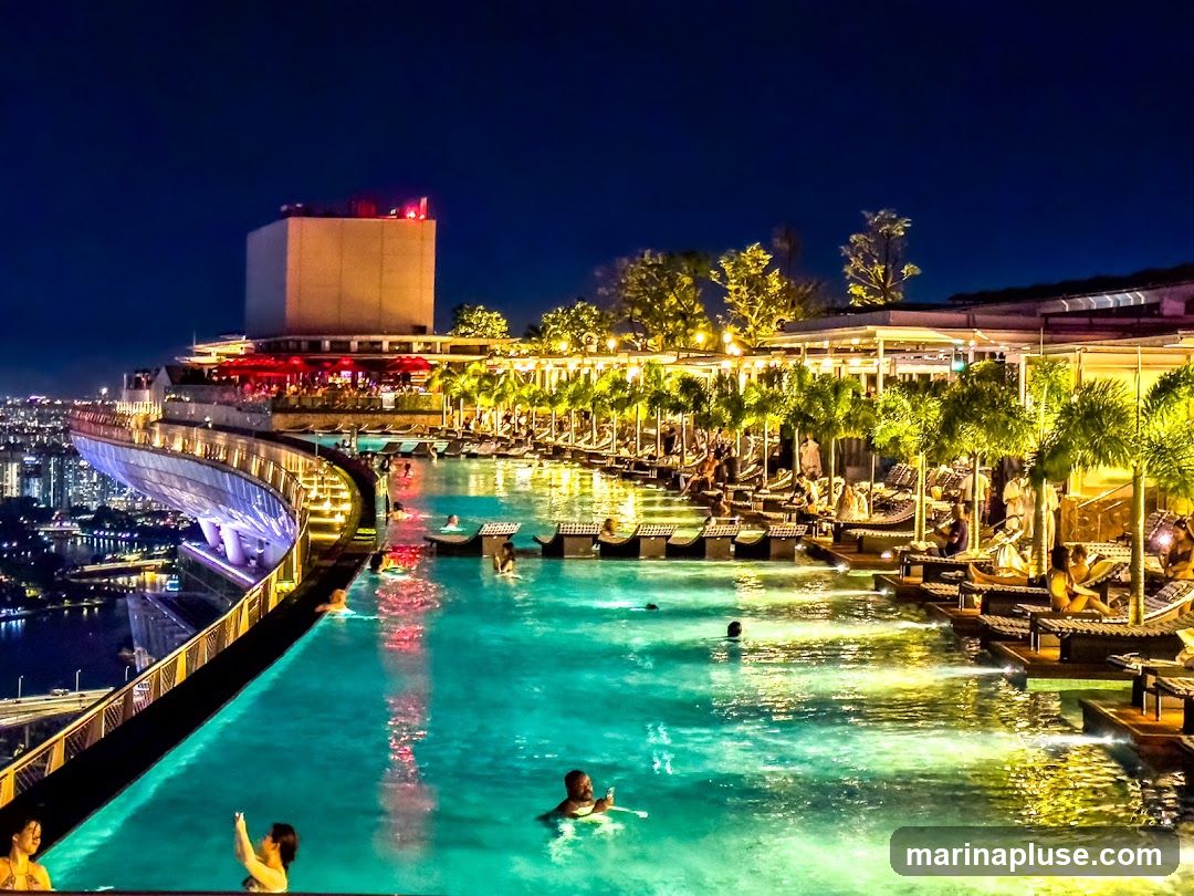 A stunning view of a rooftop infinity pool at night, with the city skyline in the background. People are swimming and relaxing on lounge chairs. The pool is illuminated with colorful lights, creating a vibrant and luxurious atmosphere. Palm trees and other tropical plants surround the pool area, adding to the resort-like feel. The architecture of the building is modern and sleek, with a large tower visible in the distance.
