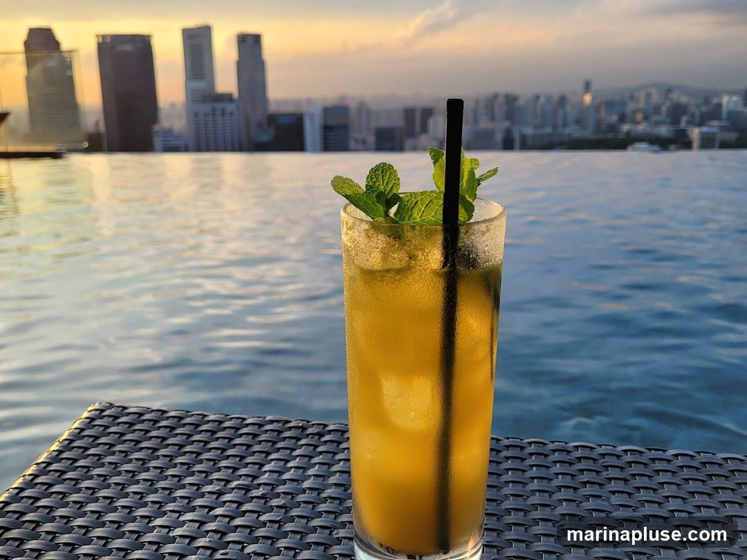 A tall glass of yellow cocktail with mint leaves and a black straw sits on a woven surface. In the background, an infinity pool reflects the sky, and a cityscape with tall buildings is visible under a sunset sky.