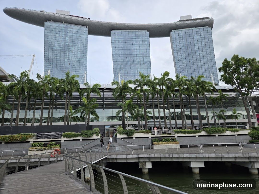 A wide shot of the Marina Bay Sands hotel in Singapore, with its distinctive boat-shaped top structure connecting three skyscrapers. In the foreground, a wooden walkway curves over a body of water, leading towards a tiered entrance with palm trees lining the facade. Several people are visible on the walkway and near the entrance. The sky is overcast.