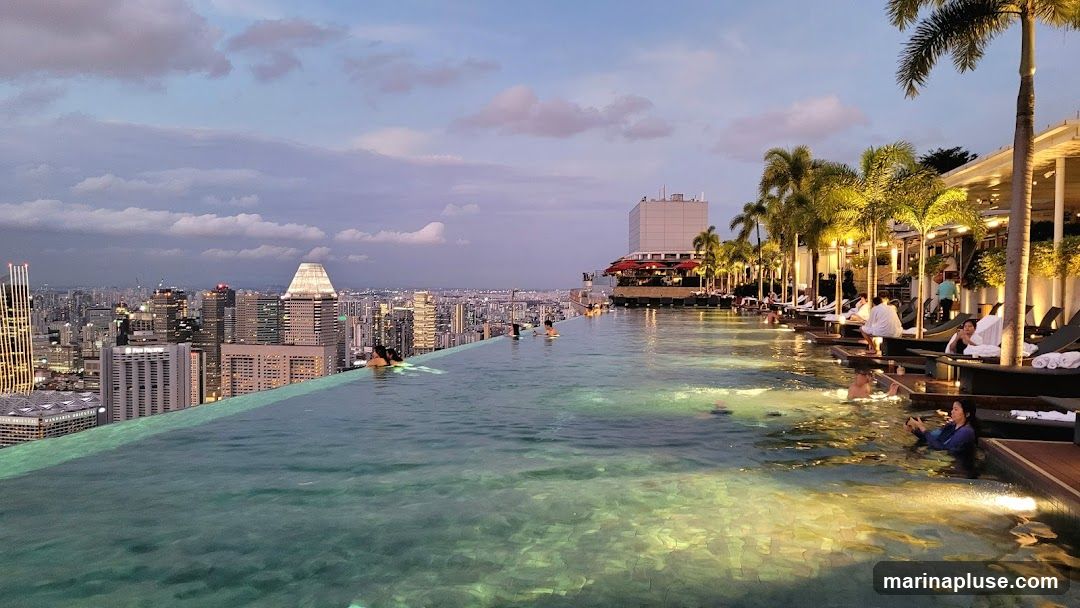 An infinity pool stretches across the foreground, with people swimming and relaxing in the water. Beyond the pool, a panoramic view of a dense cityscape is visible, illuminated by the soft light of dusk. Palm trees line the edge of the pool area, and lounge chairs are set up for patrons. The sky is a mix of soft blues and purples with scattered clouds.