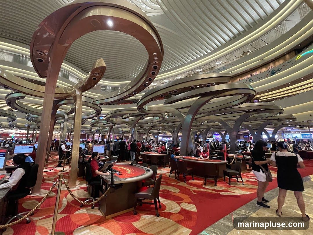 A wide shot of a bustling casino floor with numerous gaming tables and people. The architecture features large, ornate, curved structures overhead, illuminated by soft lighting. The floor is covered in a vibrant red carpet with a swirling pattern. Several roulette and card tables are visible, with dealers and patrons engaged in games. The overall atmosphere appears lively and upscale.