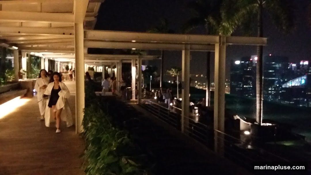 A nighttime view of a covered walkway with people walking and standing. Palm trees are visible, and in the background, a city skyline with illuminated buildings is seen. The walkway has a modern design with a sloped roof supported by pillars.