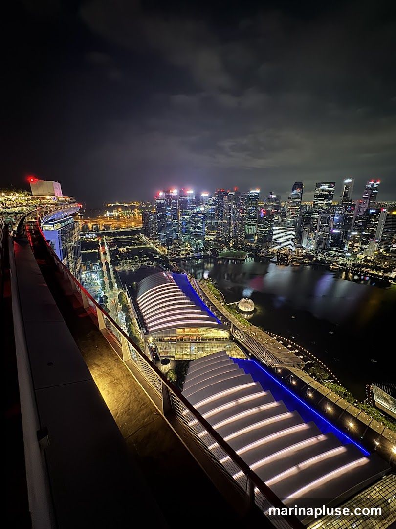 A high-angle night view of a city skyline, featuring illuminated skyscrapers and a waterfront area with modern architecture. The foreground shows a balcony or rooftop edge with railings, overlooking the cityscape. The sky is dark with some clouds.