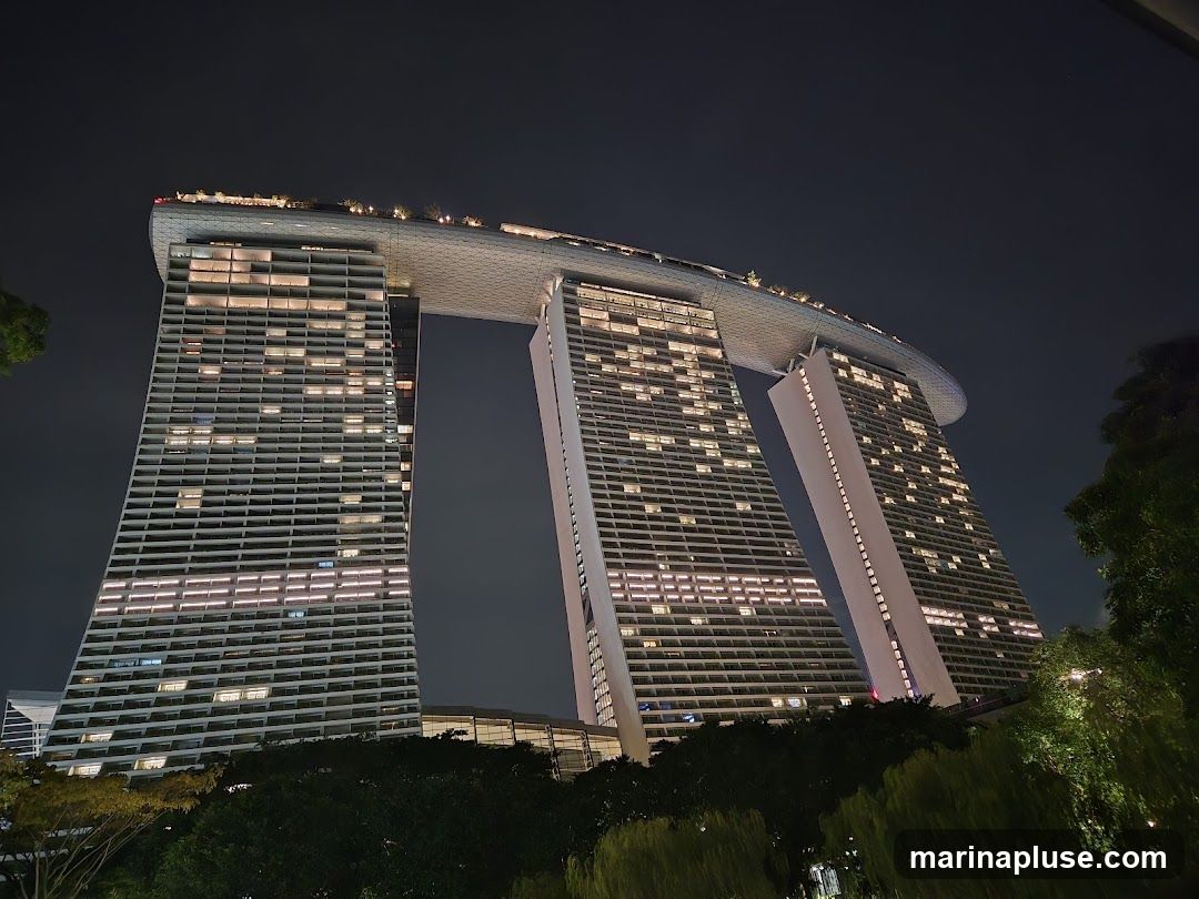 A low-angle night view of the iconic Marina Bay Sands hotel in Singapore. The three towers are illuminated, with many windows lit up, and the distinctive boat-shaped rooftop structure is visible. Trees and foliage are in the foreground, silhouetted against the dark sky.
