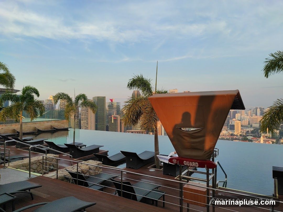 An infinity pool overlooks a sprawling cityscape at sunset. Palm trees line the pool deck, which features numerous lounge chairs. A lifeguard station with a red rescue buoy labeled 'GUARD' is visible. The sky is a soft blue with wispy clouds, and the city lights are beginning to twinkle.