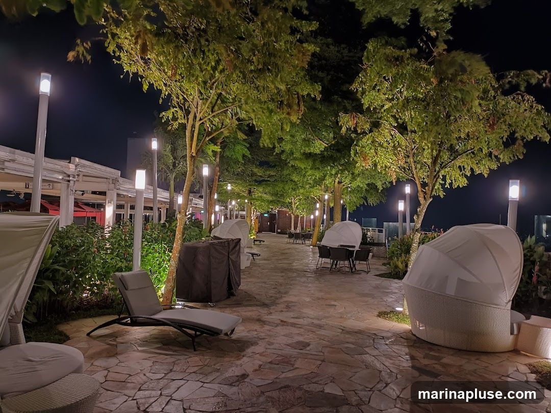 An outdoor seating area at night, illuminated by lampposts and ambient lighting. The area features stone paving, lush greenery, and trees. Several lounge chairs and tables with chairs are visible, some covered with protective canopies. The overall ambiance suggests a relaxed and upscale outdoor dining or lounge space.