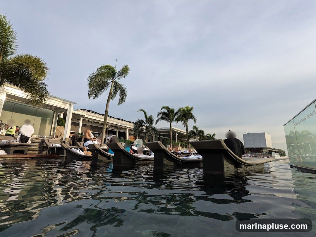 A low-angle shot of a rooftop pool with several people lounging on submerged chairs. Palm trees and modern architecture are visible in the background under a cloudy sky. A glass barrier with text reflecting the surroundings is on the right.