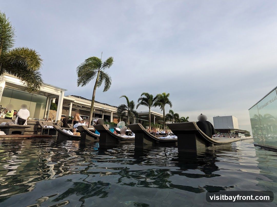 A low-angle shot of a rooftop pool with several people lounging on submerged chairs. Palm trees and modern architecture are visible in the background under a cloudy sky. A glass barrier with text reflecting the surroundings is on the right.