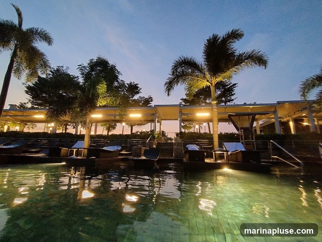 A serene view of a poolside lounge area at dusk. The water in the foreground reflects the warm glow of overhead lights and the soft hues of the twilight sky. Several lounge chairs are neatly arranged on a raised platform, some with white towels draped over them. Palm trees and other lush greenery are visible, adding to the tropical ambiance. The covered seating area with lights suggests a place for relaxation and enjoyment as evening sets in.