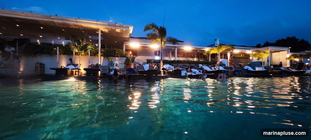 A wide shot of a resort pool area at dusk. People are relaxing on lounge chairs and in the water. The pool is illuminated, reflecting the lights from the surrounding buildings and palm trees. The architecture suggests a modern, upscale establishment with covered seating areas and a long, sleek building in the background.