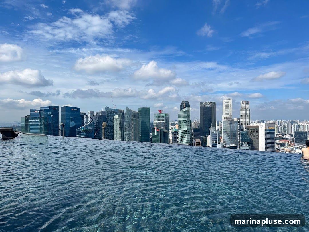 An infinity pool with rippling blue water in the foreground, overlooking a dense cityscape of modern skyscrapers under a bright blue sky with scattered white clouds. The buildings are a mix of glass and concrete, with some bearing corporate logos. A person is visible in the water on the right side of the frame.