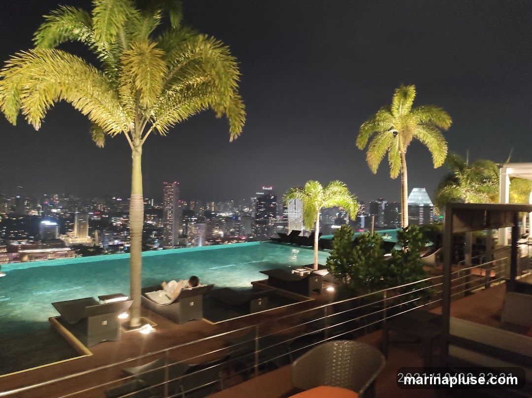 An infinity pool is illuminated at night, overlooking a sprawling cityscape. Palm trees frame the view, and lounge chairs are scattered around the pool deck. One person is relaxing on a lounge chair. The scene suggests a luxurious rooftop bar or hotel.