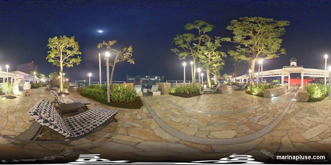 A panoramic view of a rooftop lounge area at night. The area features stone pathways, lounge chairs, trees, and ambient lighting. In the background, there is a pool area and a covered structure with seating. The sky is dark with a visible moon.