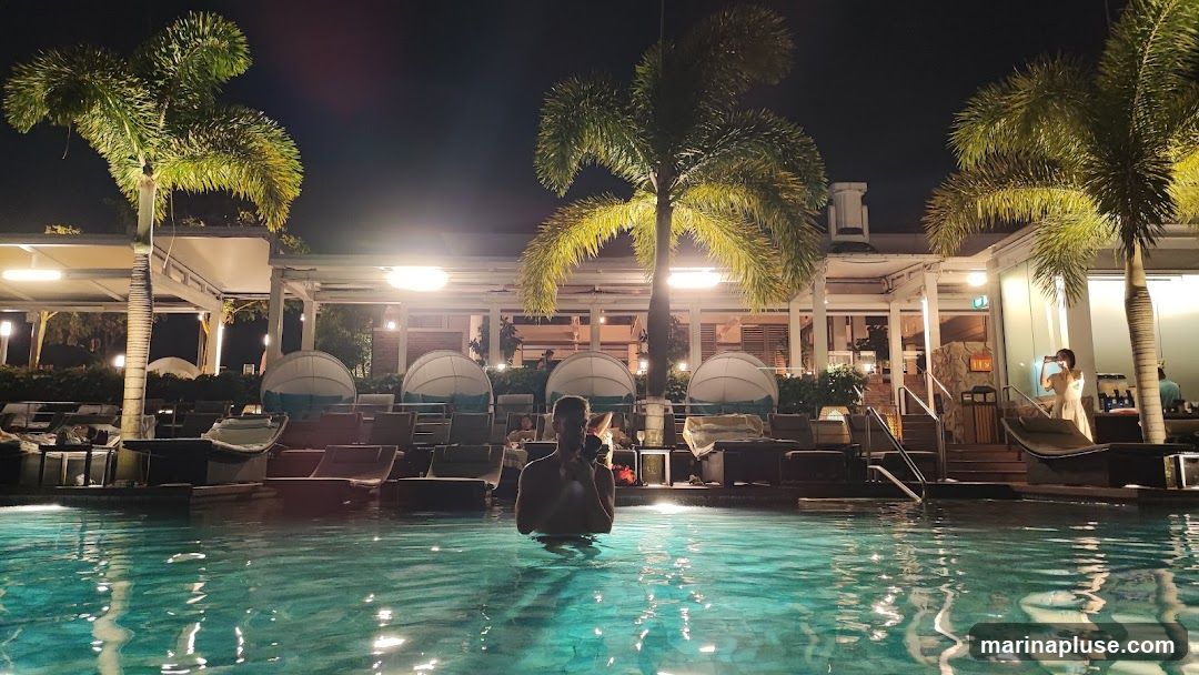 A man stands in a glowing turquoise swimming pool at night, holding a phone. Palm trees and lounge chairs with cabana-like coverings are visible around the pool. The area is lit by overhead lights, creating a relaxed ambiance. In the background, a woman in a light-colored dress is taking a photo with her phone.