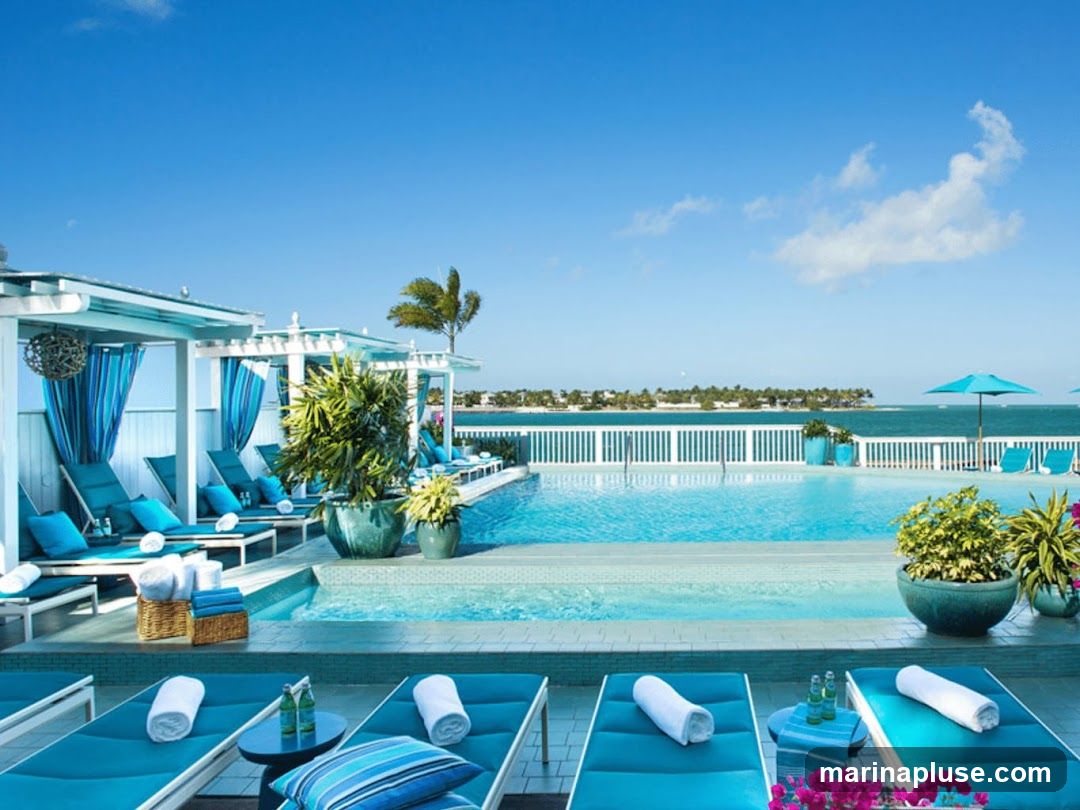 A luxurious resort with an infinity pool overlooking the ocean. Lounge chairs with blue cushions and white towels are arranged around the pool. Cabanas with blue curtains provide shade. Lush green plants in large pots add to the tropical ambiance. The sky is clear blue with a few white clouds.