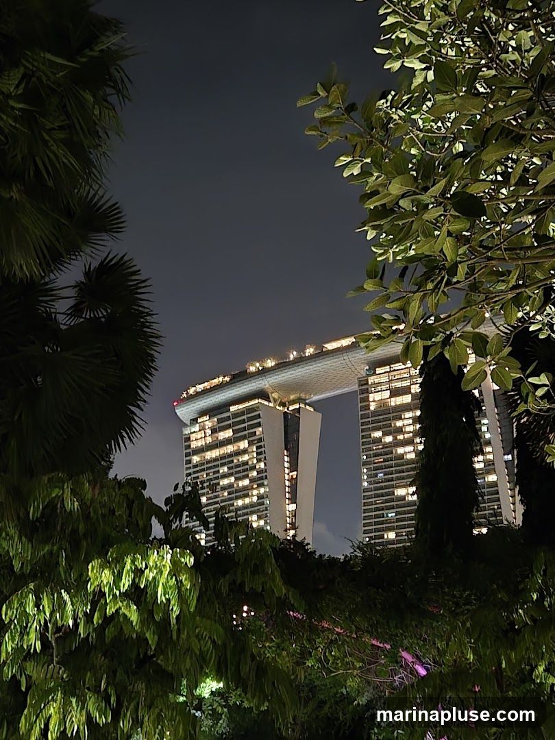 A nighttime view of the Marina Bay Sands hotel in Singapore, seen through lush green foliage. The iconic building, with its distinctive boat-shaped rooftop, is illuminated by numerous lights, creating a warm glow against the dark sky. The surrounding trees and plants frame the architectural marvel, adding a natural element to the urban landscape.