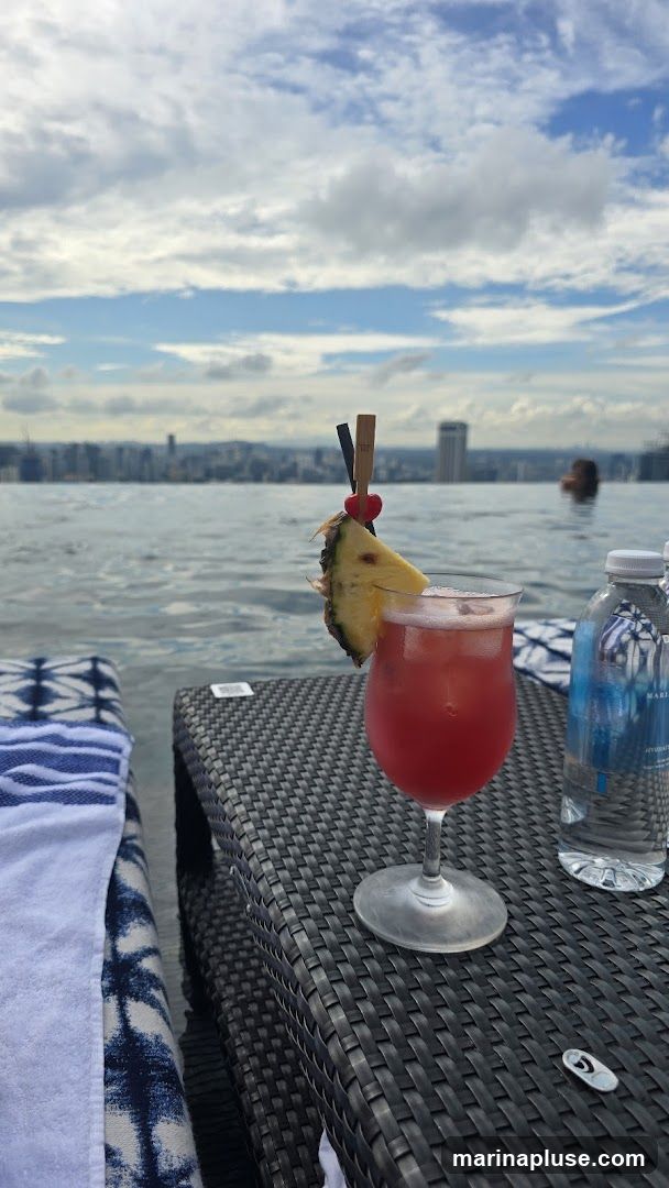 A red cocktail garnished with pineapple and a cherry sits on a woven table next to a bottle of water. In the background, an infinity pool overlooks a city skyline under a cloudy sky. A person is visible swimming in the pool.