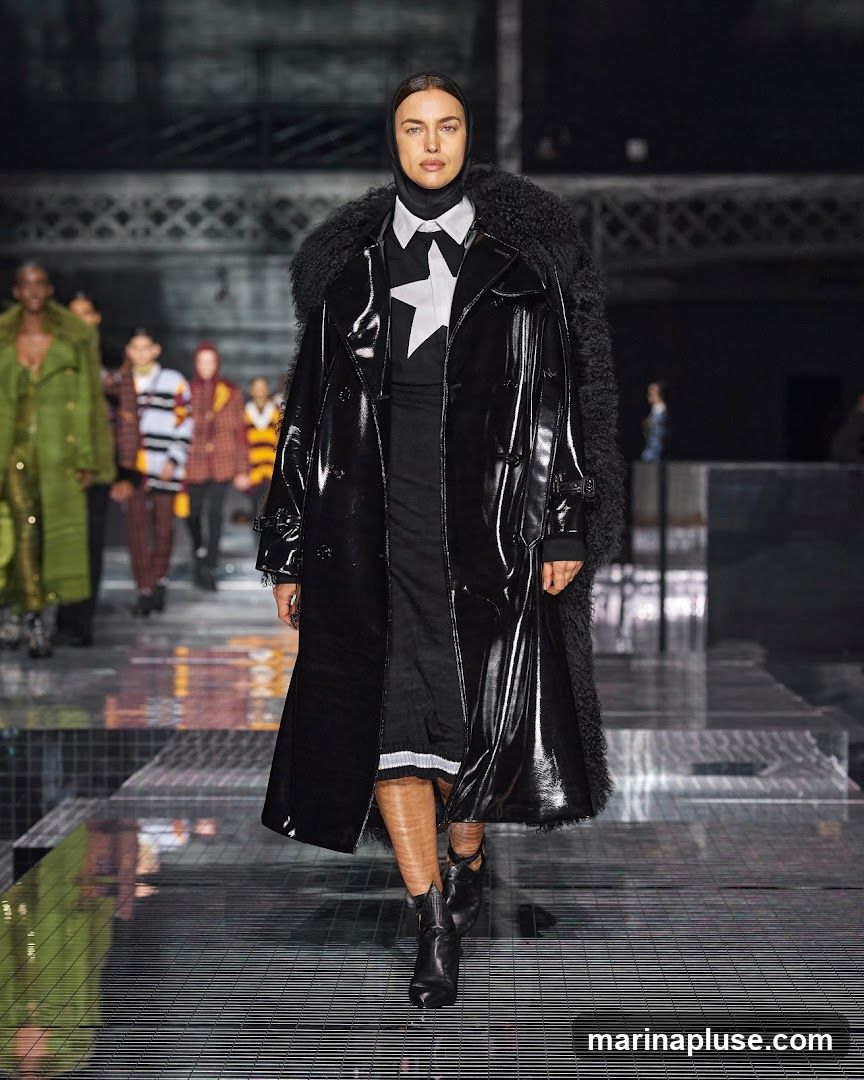 A fashion model walks down a runway wearing a black patent leather trench coat with a fur collar, a white shirt with a black star detail, and black boots. Other models are visible in the background. The runway has a reflective surface.