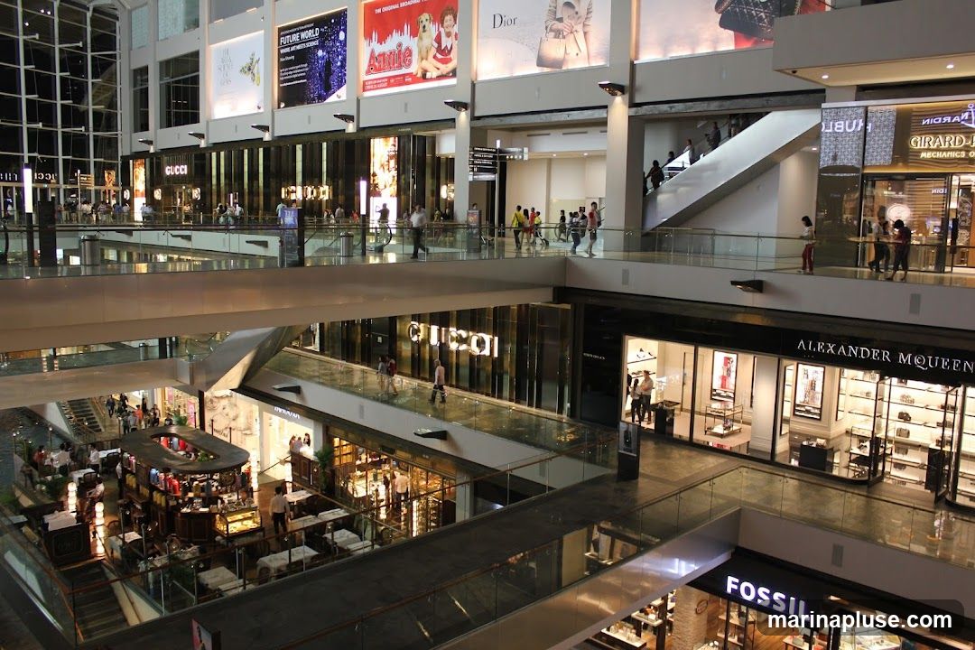 The image shows the interior of a large, multi-level shopping mall. Several walkways and escalators connect different floors. Various retail stores are visible, including GUCCI, ALEXANDER MCQUEEN, and FOSSIL. Large advertisements are displayed on the upper levels. People are seen walking around and browsing in the shops. A central area appears to be a cafe or restaurant with seating.