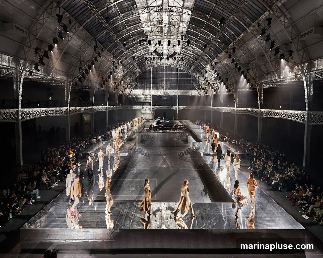 A fashion show is taking place in a large, industrial-style venue with a high, arched ceiling and exposed metal beams. A long, reflective runway stretches through the center of the space, with models walking down it. Rows of spectators are seated on either side of the runway, and spotlights illuminate the scene. A piano is visible on a raised platform at the far end of the runway.